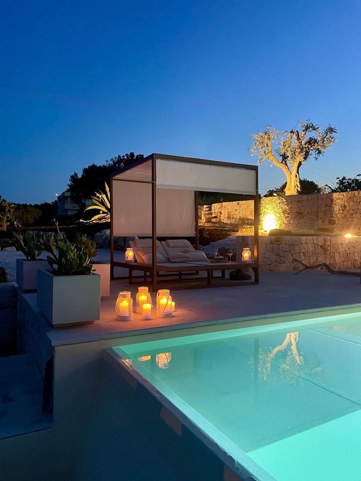 La piscina dei Trulli Bianchi al tramonto, con il baldacchino, le lanterne accese e un ulivo illuminato