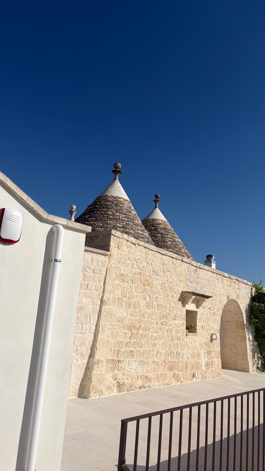 I coni dei trulli restaurati e il muro in pietra con l’arco d’ingresso, contro un cielo blu intenso